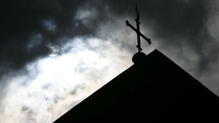 Im Gegenlicht und vor Wolken verhangenem Himmel ist die Kirchturmspitze des Doms mit Kreuz zu sehen.  Foto: Friso Gentsch/dpa