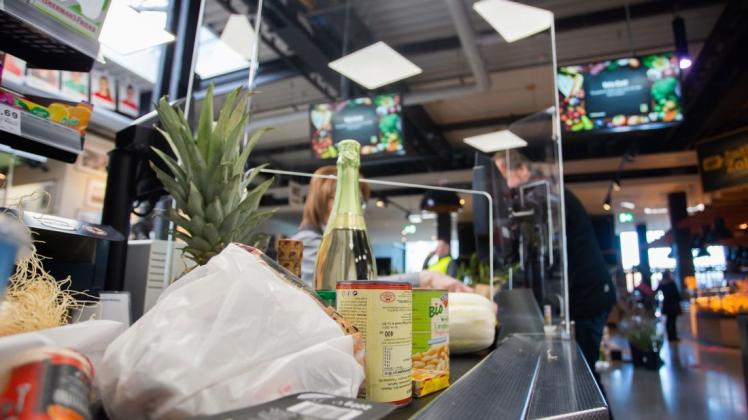 Lebensmittel stehen auf einem Förderband an einer Kasse in einem Supermarkt. Die Kassiererin wird mit einer Plexiglasscheibe, einem sogenannten "Spuckschutz", geschützt. Foto: dpa