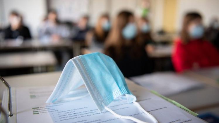 Eine Maske liegt im Unterricht auf Unterlagen, während im Hintergrund Schülerinnen und Schüler in einem Gymnasium mit Mund- und Nasenschutz zu sehen sind.