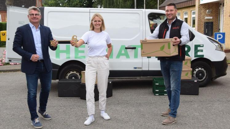 Symbolische Fahrzeugübergabe: Amtsdirektor Andreas Betz (l.) übergibt einen mächtigen Schlüssel für den Renault Traffic an Sarah Lausch von der Firma Lodeli. Diese betreibt den Online-Marktplatz Heimatware und liefert die Waren aus. Kai Lass, Geschäftsführer der Wirtschaftsfördergesellschaft des Kreises, hält einen der Pappkartons in den Händen, in denen die Produkte ausgeliefert werden. Auf dem Boden sind Styroporkisten zu sehen, in denen zu kühlende Waren transportiert werden.