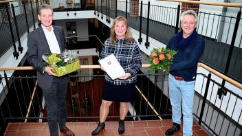 Bürgermeister Ralf Wessel (links) überreicht gemeinsam mit dem Personalratsvorsitzenden Heiko Zimmermann (rechts) Jubilarin Birgit Geerken eine Urkunde, einen Blumenstrauß und ein Geschenk.