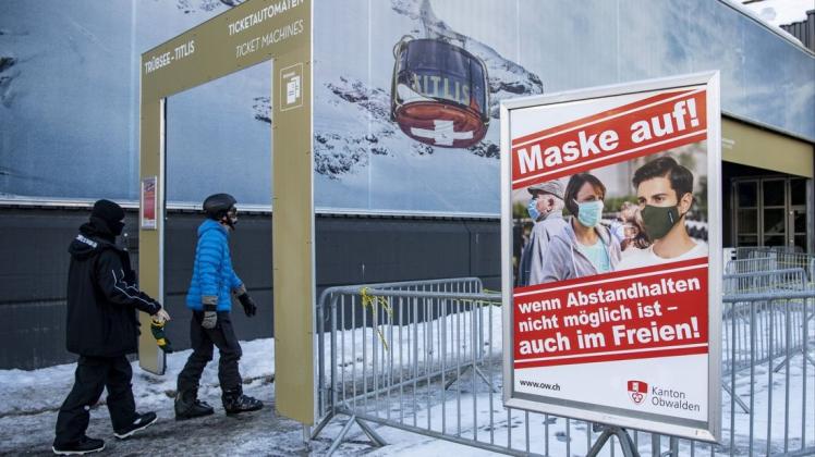 Skifahrer gehen vor der Talstation der Engelberg-Titlis-Bergbahnen durch einen Sicherheitsbereich, an dem ein Schild auf die Maskenpflicht als vorbeugende Maßnahme gegen die Verbreitung des Coronavirus hinweist.