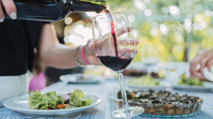 Crop unrecognizable woman pouring red wine from bottle into glass while standing at table during outdoor lunch in garden
