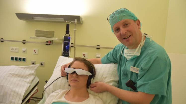 Narkose-Arzt Dr. Günter Riese (rechts) stellt seiner Patientin vor der OP die audiovisuelle Brille nach Seh- und Hörstärke ein – mit einer Komödie geht nun die Wartezeit vor der Operation ein wenig entspannter vorbei. 