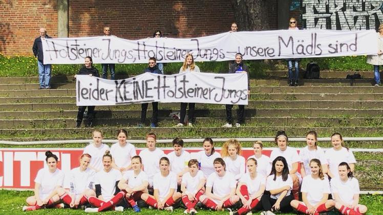 „Holsteiner Jungs, Holsteiner Jungs – unsere Mädls sind leider keine Holsteiner Jungs“ steht auf einem Spruchband. Das Frauenteam sitzt davor auf dem Rasen.