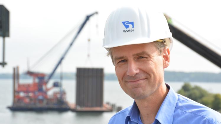 Gut im Zeitplan: WSA-Projektleiter Matthias Baasch lächelt zufrieden in die Kamera. Im Hintergrund hängt ein Segment an den Seilen des Schwimmkrans. 