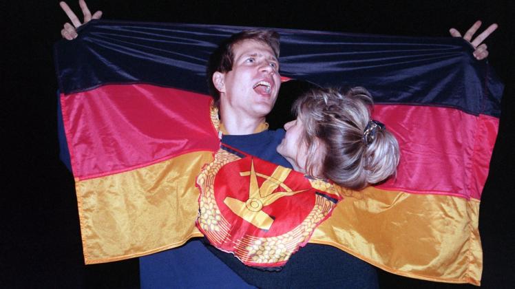 03.10.1990, Berlin: Ein jubelnden Paar hat sich am Tag der Deutschen Einheit in einer DDR-Fahne dort Platz geschaffen, wo früher das Emblem Hammer und Sichel war - und feierte frenetisch den historischen Anlaß.