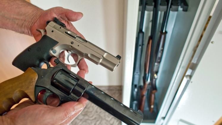 Ein Sportschütze zeigt eine Pistole und einen Revolver vor einem Waffenschrank mit drei Gewehren in einem privaten Haushalt. 