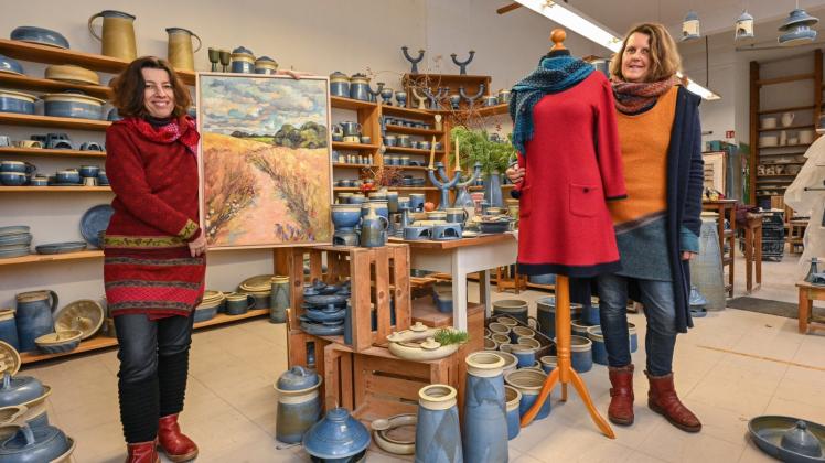 Christine Radecke (Künstlerin mit einem Gemälde), steht zusammen mit Anke Müller, Walkkleidung Wollbrunnen, in der Keramikwerkstatt von Anne Schattka-Steinbruch auf dem Gelände des Kunsthandwerkerhofes. 