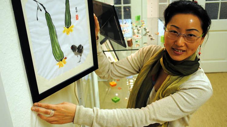 Yibo Beck hängt eine Arbeit mit Tuschemalerei auf Reispapier für ihre Ausstellung in der Kreisvolkshochschule auf. Im Hintergrund eine Vitrine mit Jade-Schmuck. 