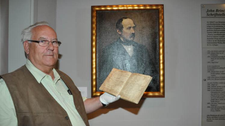 Vor einem Porträt von John Brinckman zeigt Dieter Kölpien das Kochbuch von Elise Brinckman. 