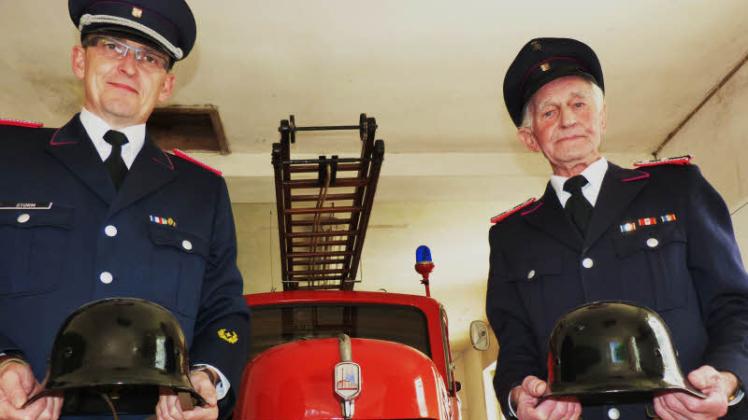 Gemeindewehrführer Thomas Storm (46, links) und Oberfeuerwehrmann Dirck Boyens (86) stehen vor dem historischen Fahrzeug namens  Minna. In den Händen halten sie die alten Schutzhelme aus Stahl.   