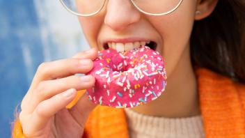 Positive cropped unrecognizable woman in knitted warm cardigan eating tasty pink sprinkled donut DavidMunoz_LucilOrange_