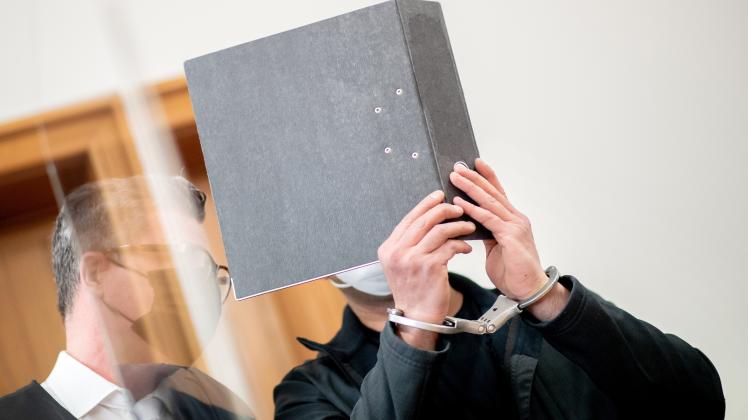 Der angeklagte 34-Jährige steht zu Prozessbeginn mit Handfesseln in einem Saal im Landgericht Oldenburg und bedeckt sein Gesicht mit einem Aktenordner, während er mit seinem Anwalt Detlev Otto Binder spricht.