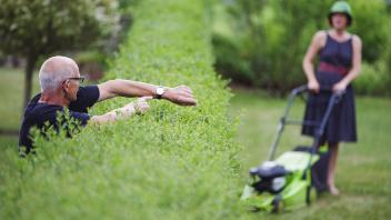 Ein Mann zeigt an einer Hecke auf seine Uhr waehrend seine Nachbarin den Rasen maeht. Martinroda, 28.07.2013. Model Rel