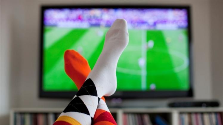 Überschlagene Beine vor einem Fernsehgerät, auf dem ein Fußballspiel übertragen wird. 