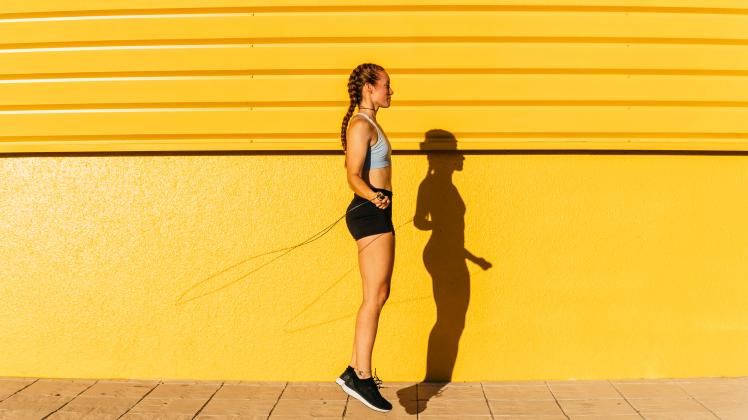 Young sportswoman using rope while skipping by yellow wall during sunny day model released Symbolfoto MGRF00441