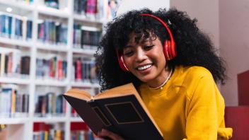Stylish happy young ethnic female student with long dark curly hair in trendy outfit and headphones on neck smiling whil