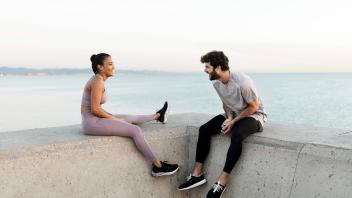 Cheerful young bearded sportsman speaking with content ethnic female athlete while resting against endless sea and looki