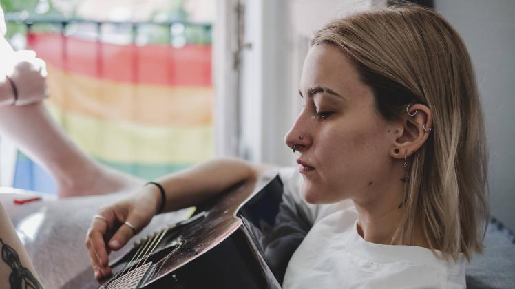 Young blonde woman with face piercings playing guitar sitting on bed near big windows at home Copyright: xDanielxGonzale