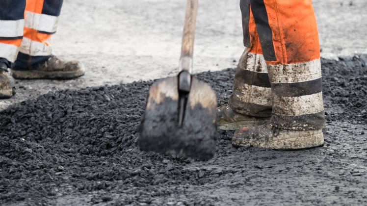 Bauarbeiterbeine in Arbeitskleidung mit einer Schaufel stehen im warmen Asphalt Straßenbau *** Construction worker legs