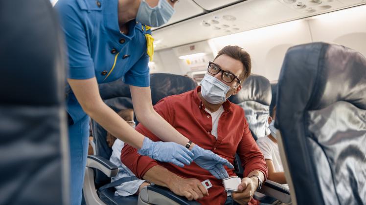 Man in protective face mask looking at female flight attendant helping him to adjust and tight a seatbelt on an airplane