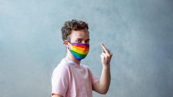 Transgender in protective mask with LGBT rainbow pattern looking at camera and showing obscene middle finger gesture on
