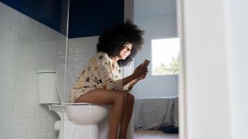 Smiling woman using smart phone while sitting on toilet seat at home model released Symbolfoto property released JCCMF03