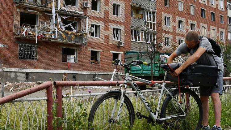 Ein Mann in Slowjansk zurrt einen Koffer auf seinem Fahrrad fest. Das Wohnhaus hinter ihm wurde bei einem nächtlichen Raketeneinschlag beschädigt. Foto: Andriy Andriyenko/AP/dpa