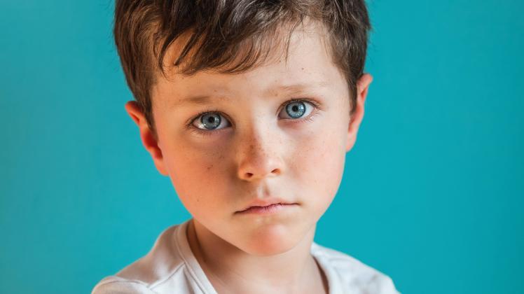 Headshot of upset boy with brown hair looking at camera with upset face expression while standing in light studio on gre