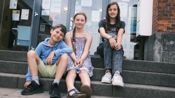 Die drei Viertklässler Emmy, Ilyas und Carolin sitzen vor ihrer Schule.