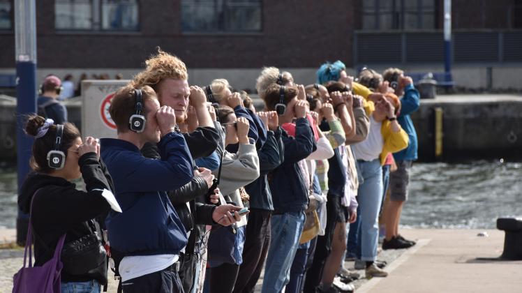 Menschen mit Kopfhörern stehen am Alten Hafen und schauen aufs Wasser - die Hände zu einem Fernglas geformt.