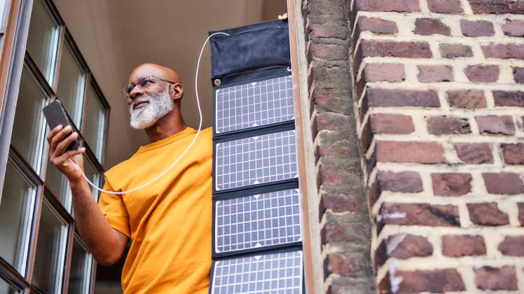 Man with smart phone and solar panel standing by window at home model released, Symbolfoto property released, FMKF07435