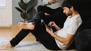 Bearded barefoot man leaning on sofa and scrolling tablet near cropped unrecognizable pregnant woman using cellphone in
