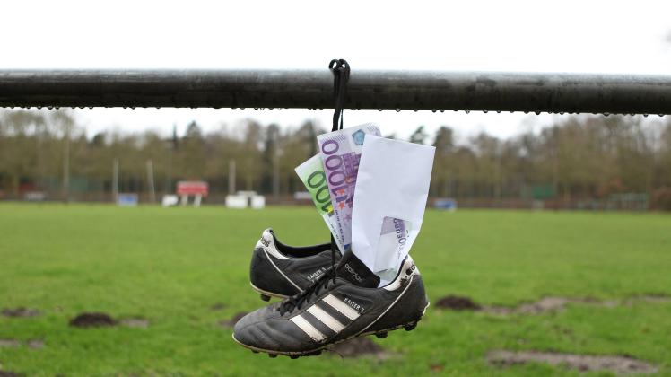 Ein Briefumschlag mit Geldscheinen steckt in Fußballschuhen, die an der Balustrade eines Sportplatzes hängen. Symbolbild