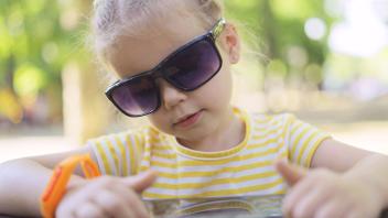 Kleines Mädchen mit Sonnenbrille lernt Fremdsprache durch Wiederholung von Wörtern aus dem Handy. Close-up-Porträt von K