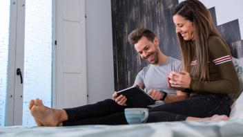 Full body of positive couple surfing tablet while sitting together on comfortable bed with cups of coffee in light bedro