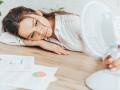 exhausted businesswoman blowing at herself with electric fan while lying on table with documents