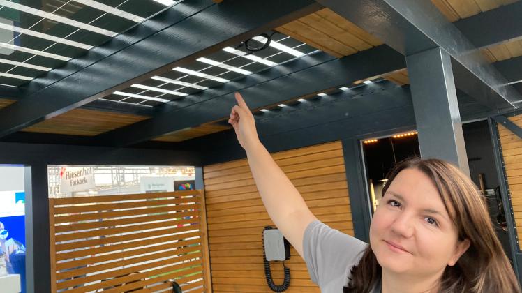 Jana Ploß vom Holzbauzentrum Nord in Kiel weist auf den in Halle 8 aufgebauten Carport mit transparenten Photovoltaik-Panelen und Wallbox zum Aufladen des Elektroautos.