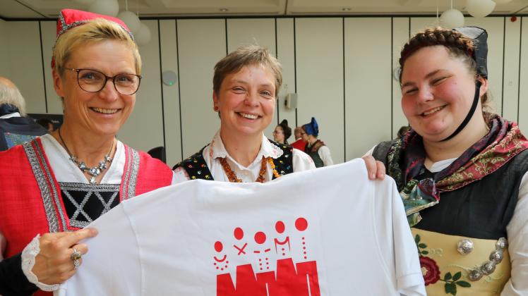 Anna Czyttrich (rechts), Katrin Rathjen und Wiebke Schweitzer-Bösenberg vom Landestrachten- und Volkstanzverband Schleswig-Holstein präsentieren das neue Logo auf einem Shirt.
