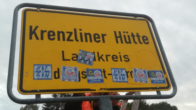 Gemeindearbeiter entfernen die Aufkleber auf dem Ortseingangsschild von Krenzliner Hütte.