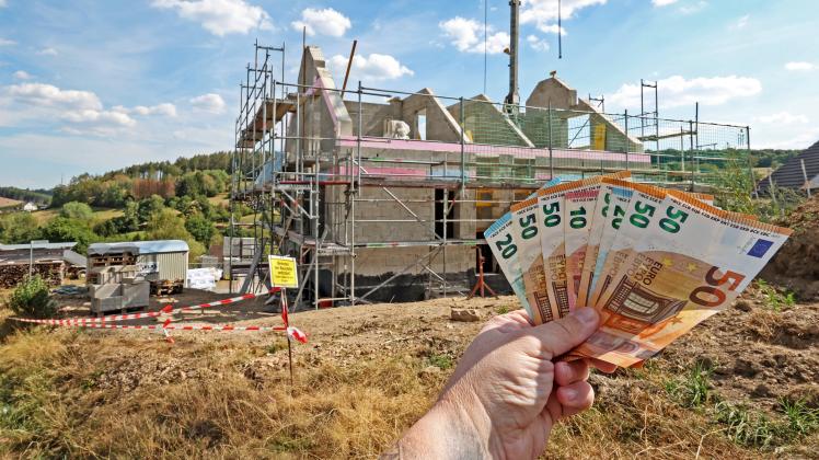 Bauen wird teurer. Wer kann zukuenftig noch bauen? Hand mit Eurogeldscheinen vor einem Neubau. Wohnungsbau, Hausbau im S