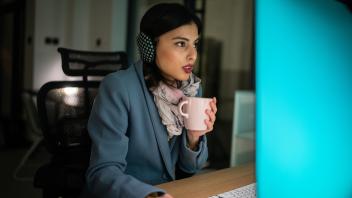 Woman in warm clothing sitting in freezing office and working on computer with hot coffee cup in hands