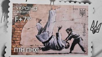 A photograph taken on February 24, 2023 shows a stamp reproducing a drawing of the famous British artist Banksy during their presentation at the main post office in Kyiv, on the first anniversary of the Russian invasion of Ukraine. - Residents of Kiev flocked to the main post office of the capital to get a new stamp reproducing a drawing of the famous British artist Banksy made on the wall of a destroyed building in Borodyanka. The work depicts a child judoka throwing a man also in judo clothing to the ground. On the bottom of the stamp was added the inscription "Fuck yourself Putin", in reference to the judo practiced by the Russian president. (Photo by Sergei SUPINSKY / AFP) / RESTRICTED TO EDITORIAL USE - MANDATORY MENTION OF THE ARTIST UPON PUBLICATION - TO ILLUSTRATE THE EVENT AS SPECIFIED IN THE CAPTION