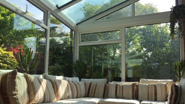 Image of rectangular white UPVC conservatory lean-to glass room with sunny garden views, large double-glazed windows and glass roof with self-cleaning panes / L-shaped corner sofa settle conservatory furniture, coffee table and television TV remote contro