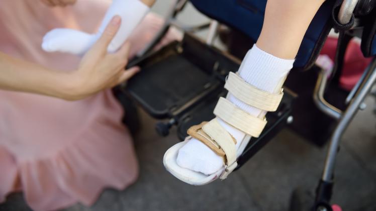 Mother puts on orthosis her daughter legs. Disabled girl sitting on a wheelchair. Child cerebral palsy.