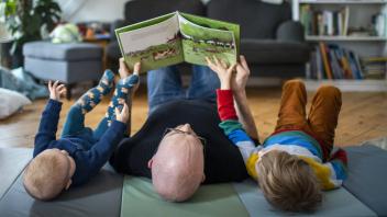 Symbolfoto: Ein Vater liest mit seinen beiden Kindern auf dem Boden liegend ein Buch. Berlin, 02.02.2021. Berlin Deutsch