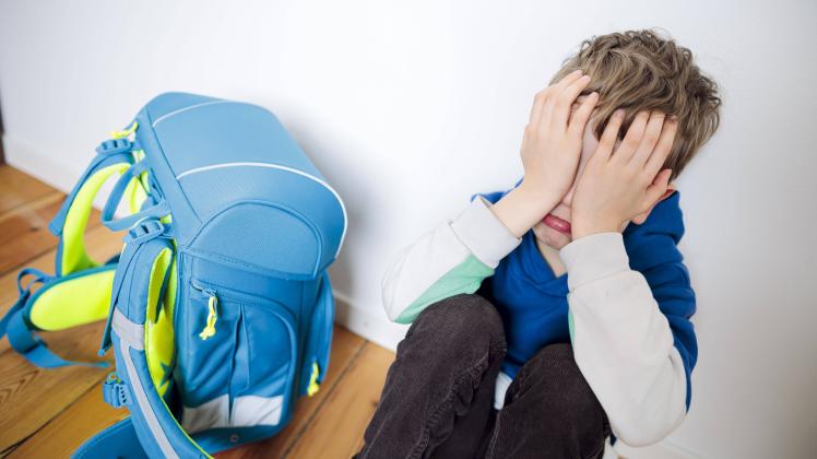 Symbolfoto zum Thema Angst vor der Schule. Ein Junge sitzt traurig in seinem Kinderzimmer neben seinem Schulranzen und v