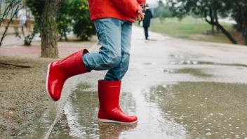 Adorable joyful child in red raincoat and rubber boots having fun jumping in puddle on street in park in gray day, Model