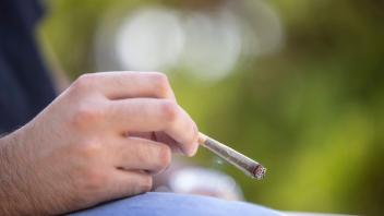 Detail of man holding a lit CBD pre-roll joint, cigarette with smoke rising sitting outdoors PUBLICATIONxNOTxINxUSA Copy